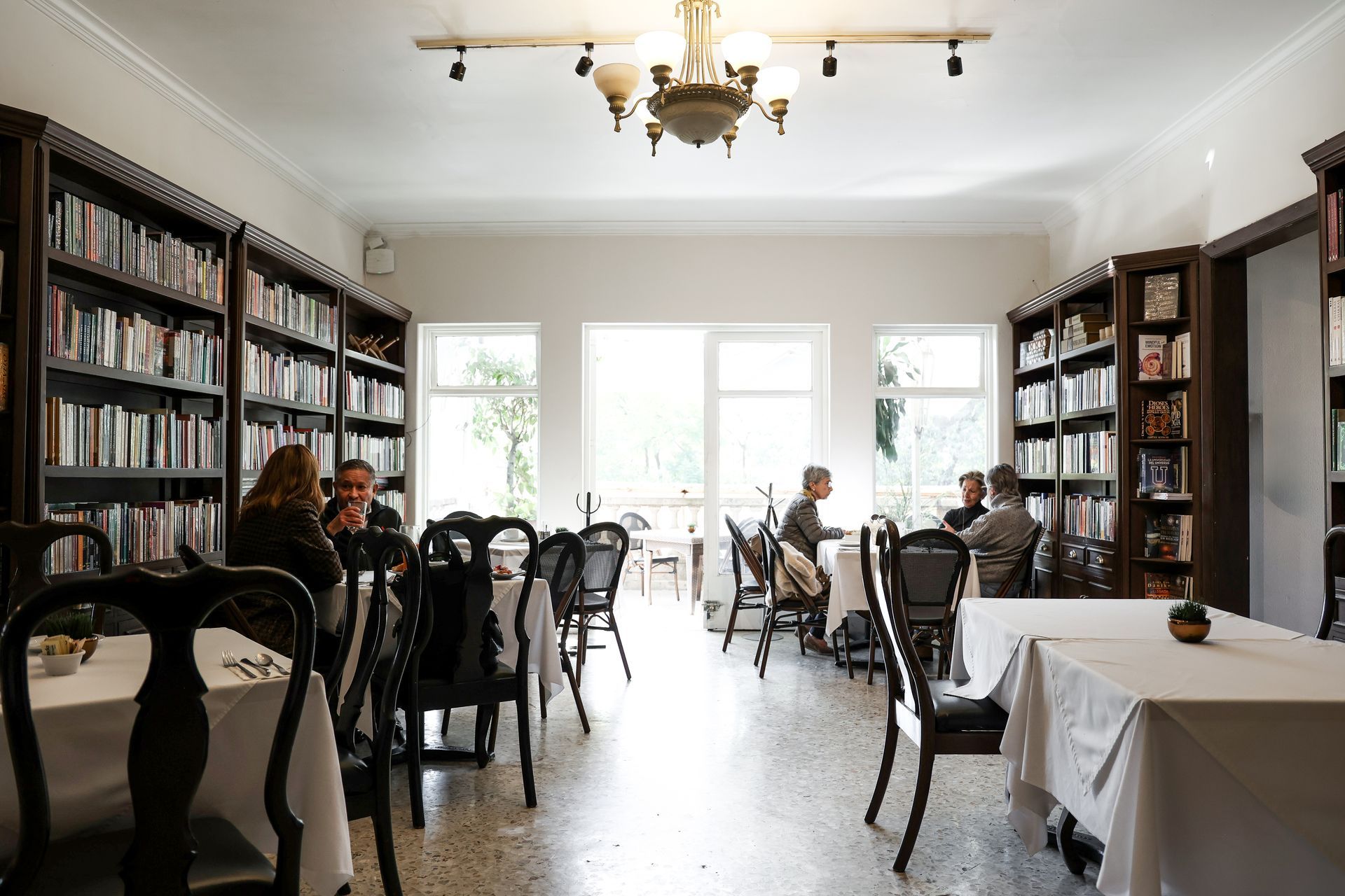Comedor luminoso con estanterías, mesas y gente sentada cerca de grandes ventanales.
