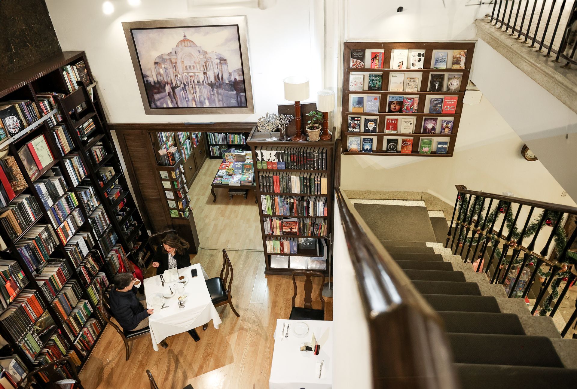 Estanterías y mesas de exposición en una acogedora librería con escalera y cuadros enmarcados en la pared.