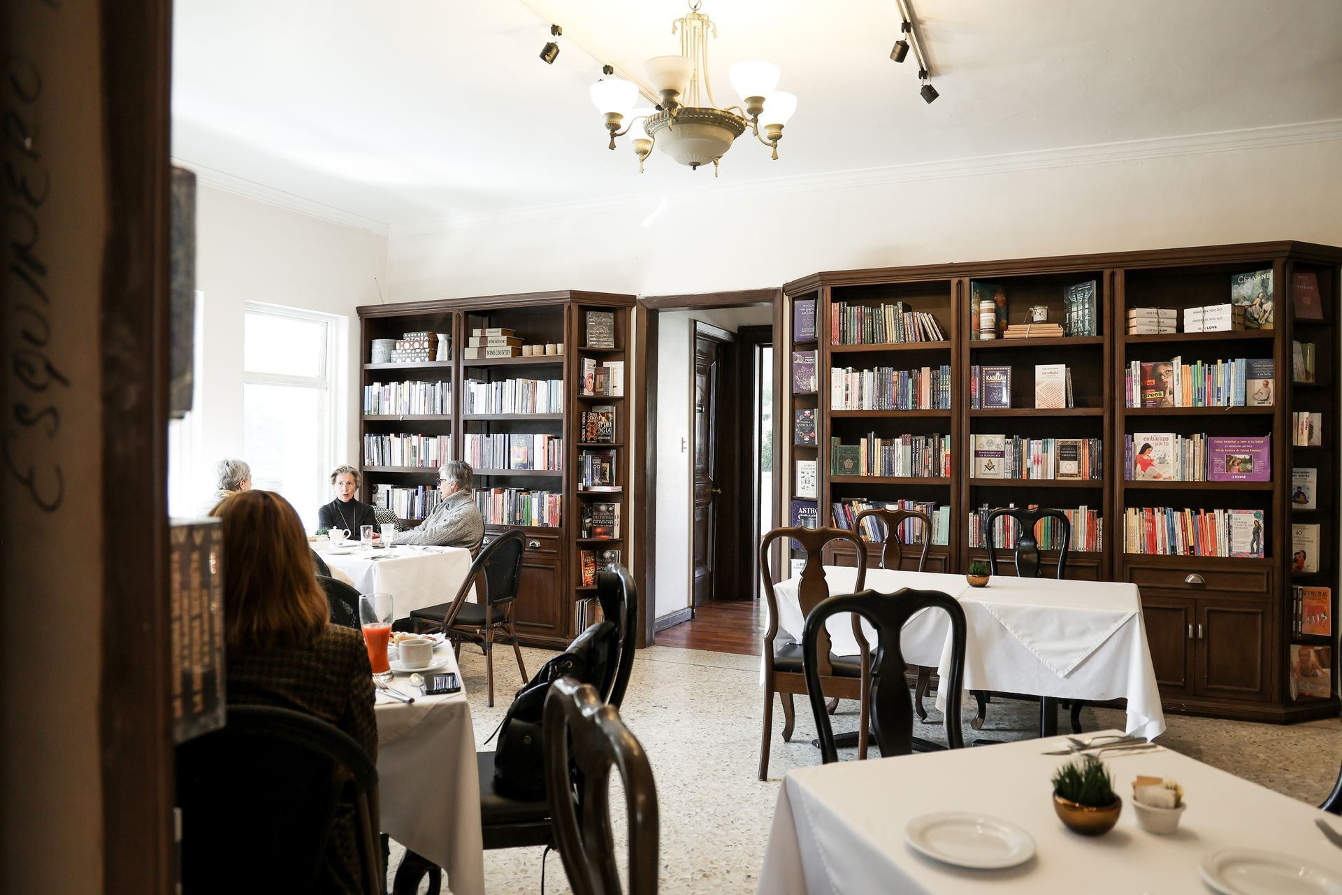 Comedor-cafetería con estanterías repletas de libros, mesas, sillas y una lámpara de araña.