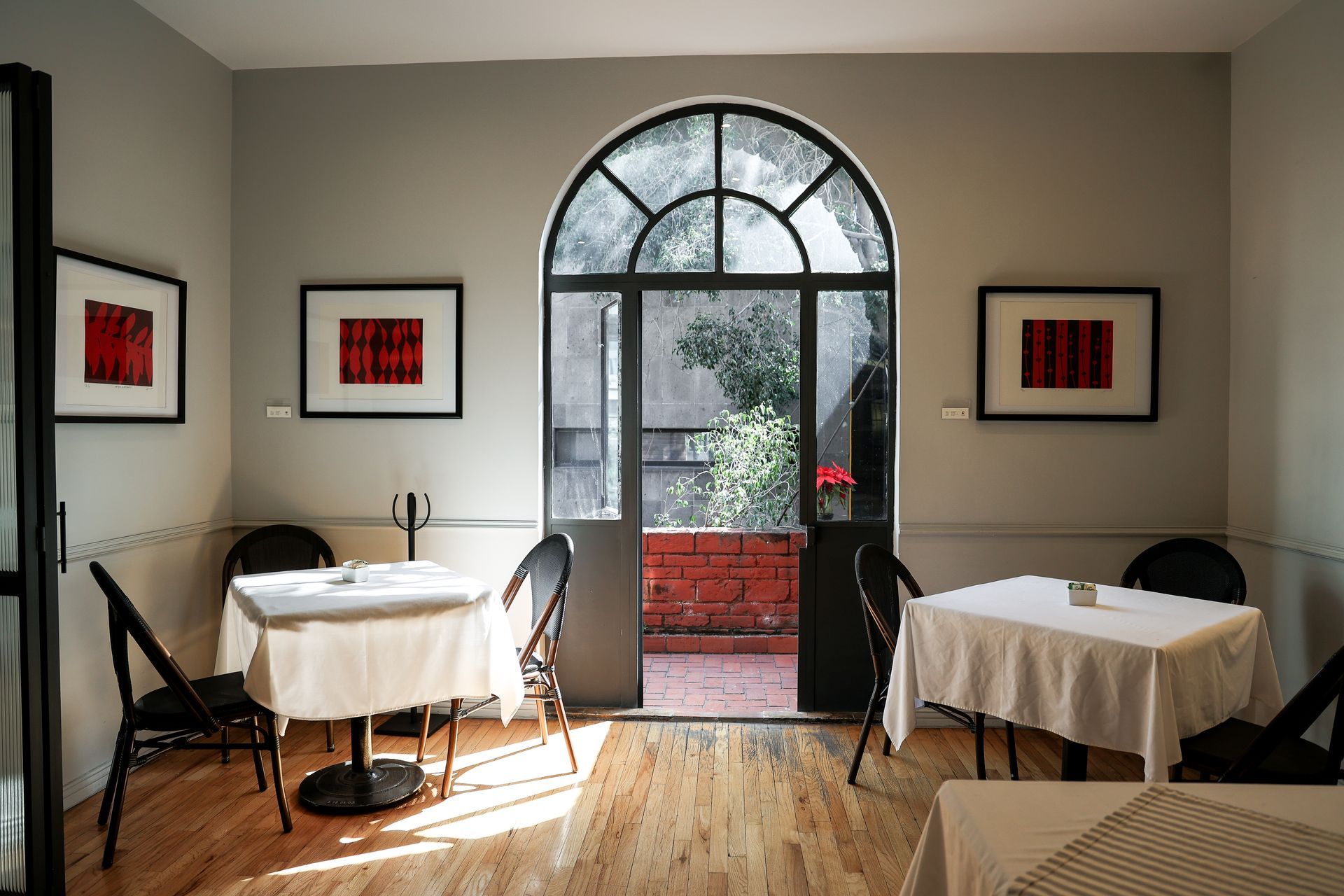 Pequeño comedor tipo cafetería con dos mesas, puerta de cristal arqueada y un cuadro rojo enmarcado.