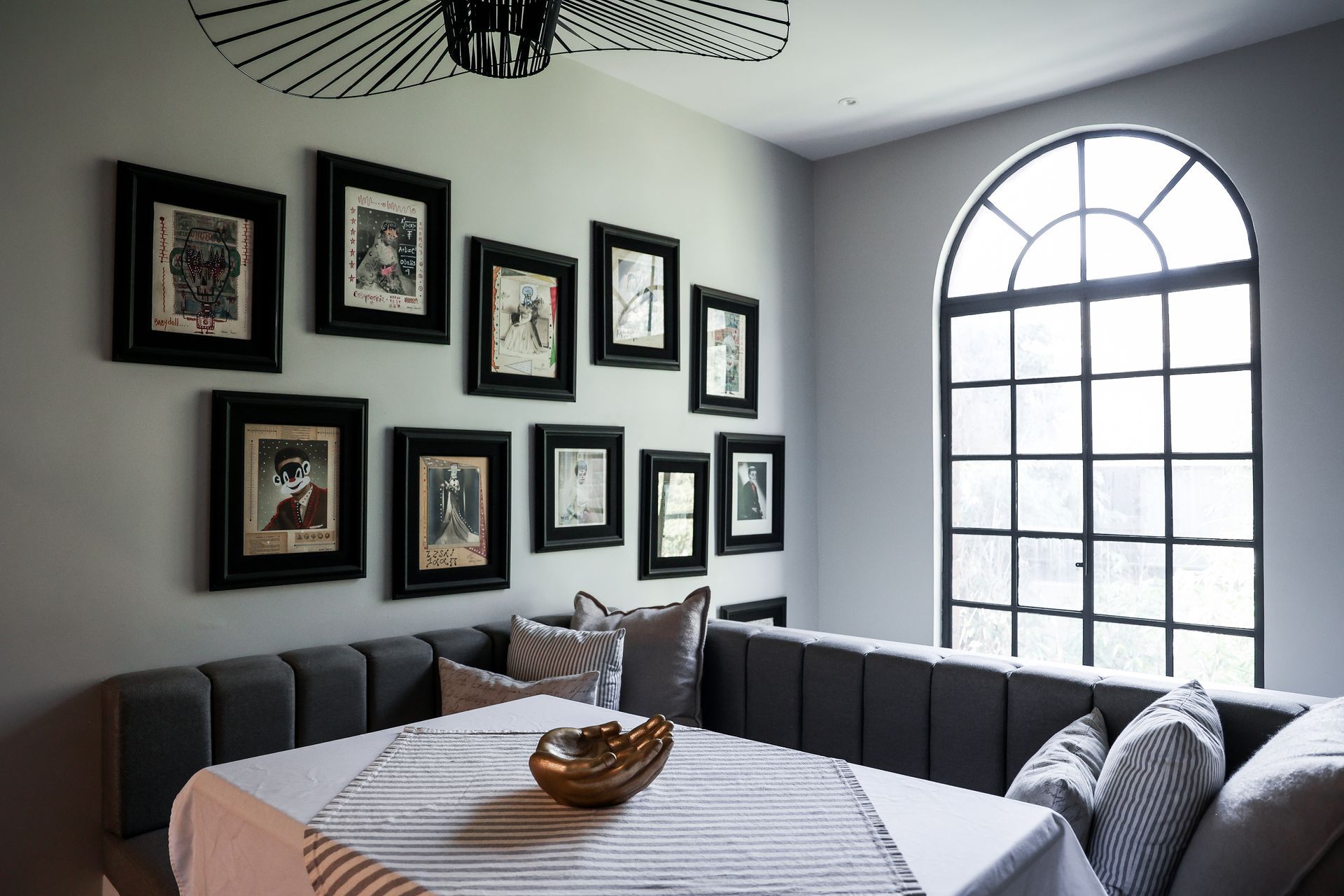Acogedor rincón de comedor con asientos tipo banqueta grises, cuadros enmarcados en la pared y una gran ventana arqueada.