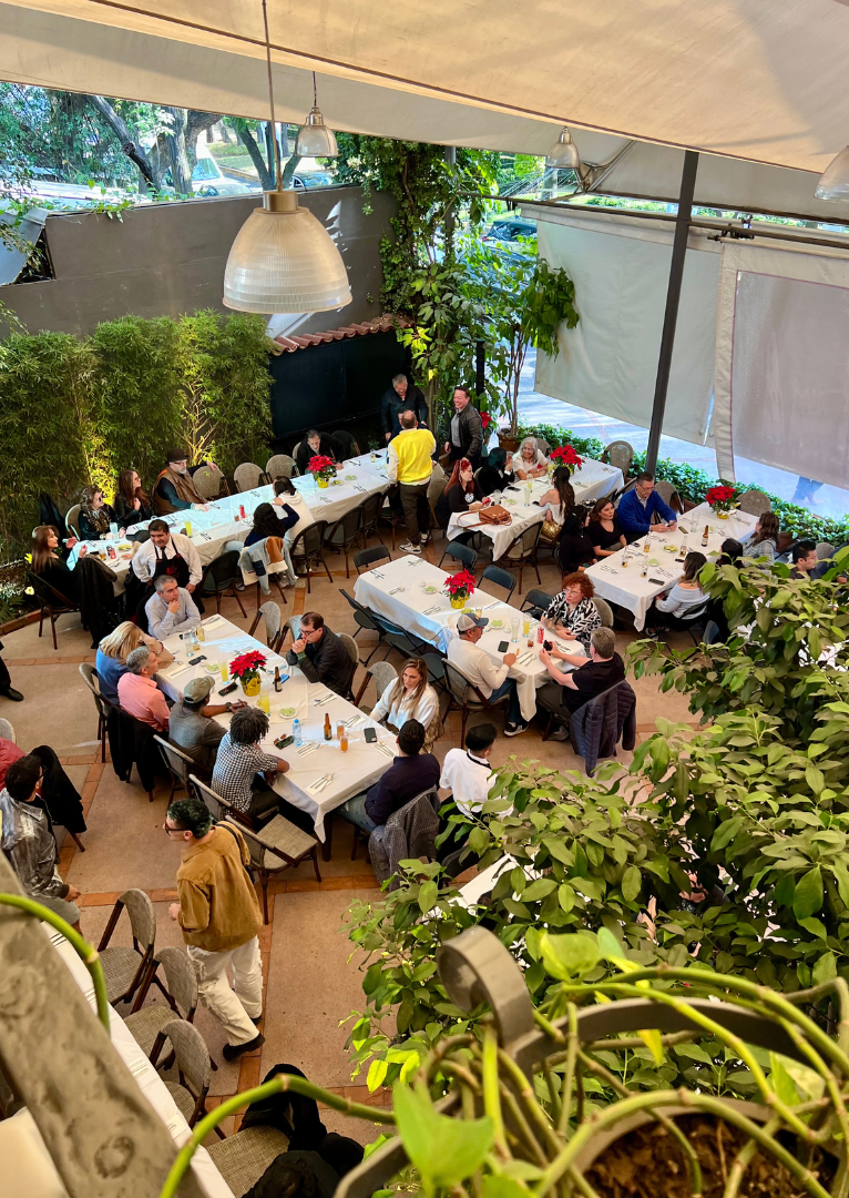 Gente sentada en largas mesas en una luminosa cafetería con jardín interior, plantas exuberantes y luces colgantes.