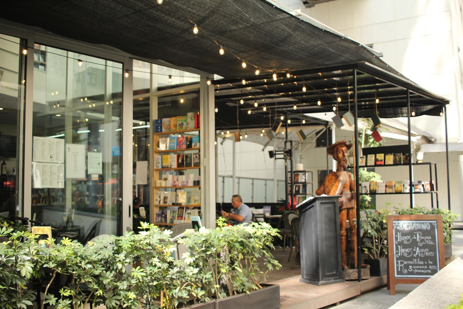 Cafetería al aire libre con toldo negro, luces colgantes, plantas, estanterías y una persona de pie junto al mostrador.