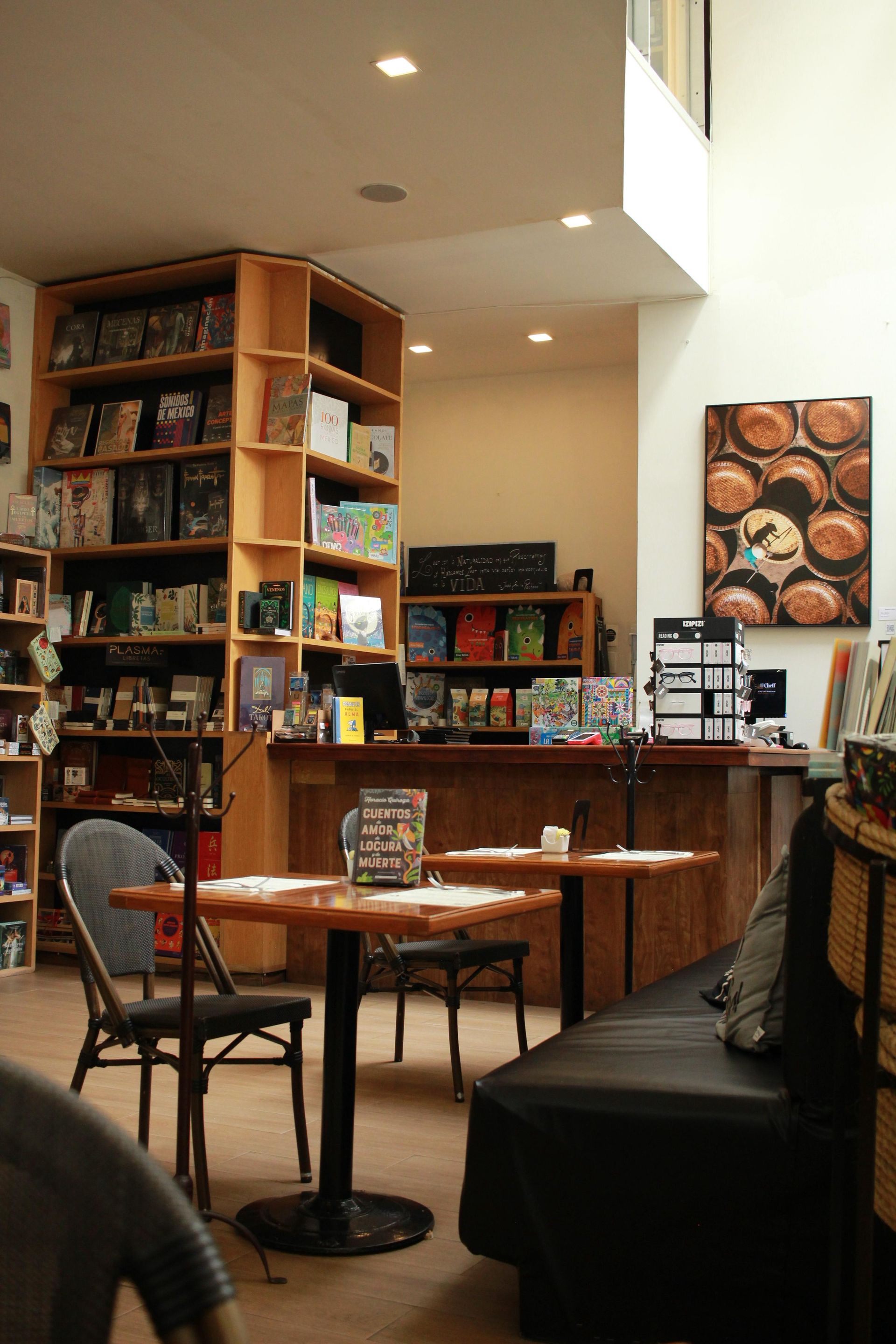 Interior acogedor de una cafetería con mesas de madera, estanterías con libros y otros artículos, y una barra en la parte trasera.