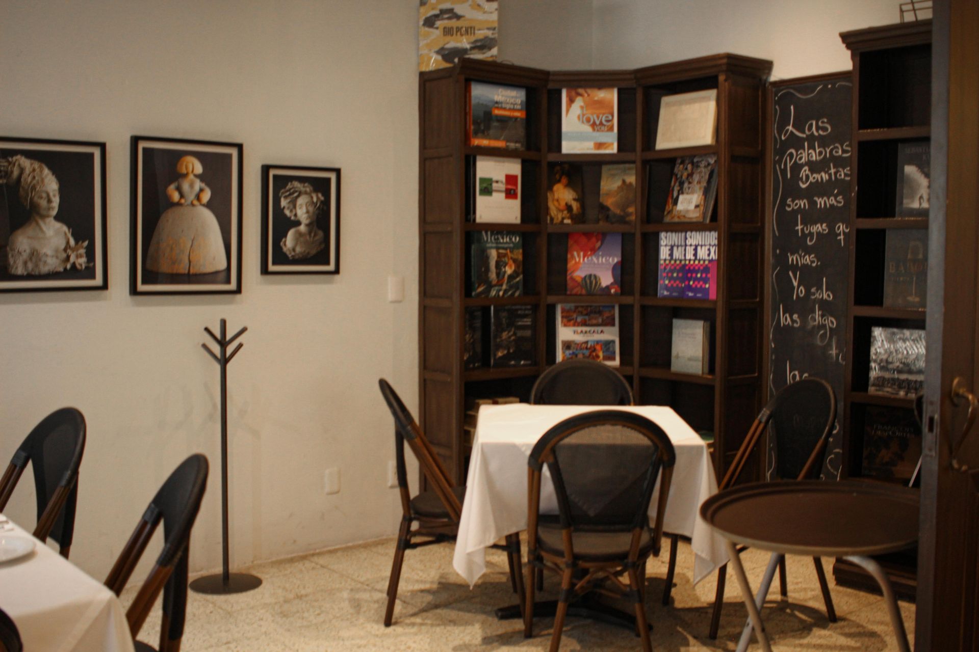 Acogedor rincón de lectura en una cafetería con retratos enmarcados, estanterías y mesas con sillas negras.
