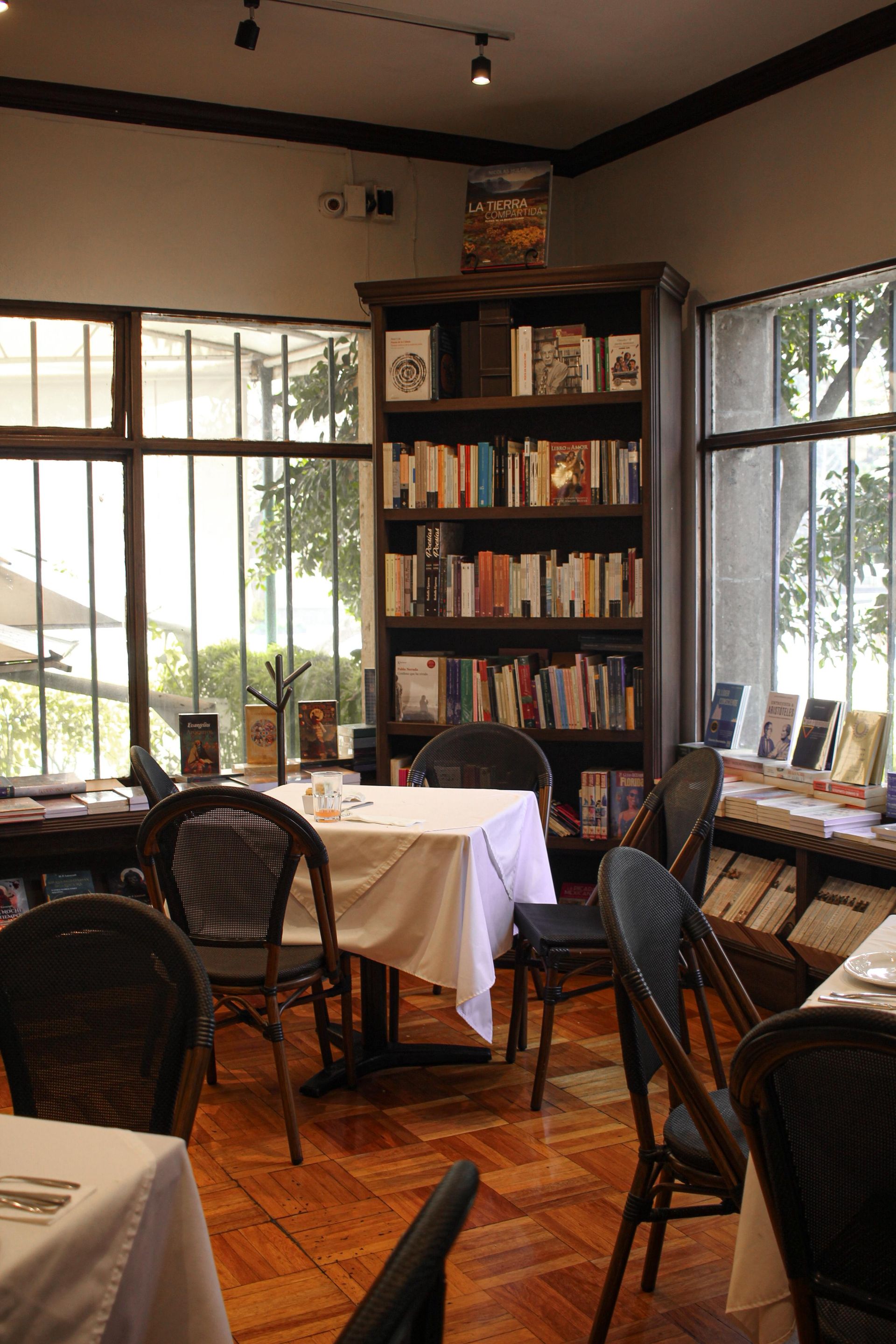 Acogedora cafetería con sillas de mimbre, manteles blancos, estanterías y grandes ventanales con vistas a la vegetación.