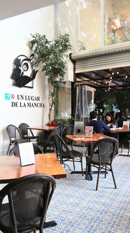 Terraza de café al aire libre con sillas negras, suelo de mosaico, árbol en maceta y cartel que dice “Un lugar de la Mancha”.