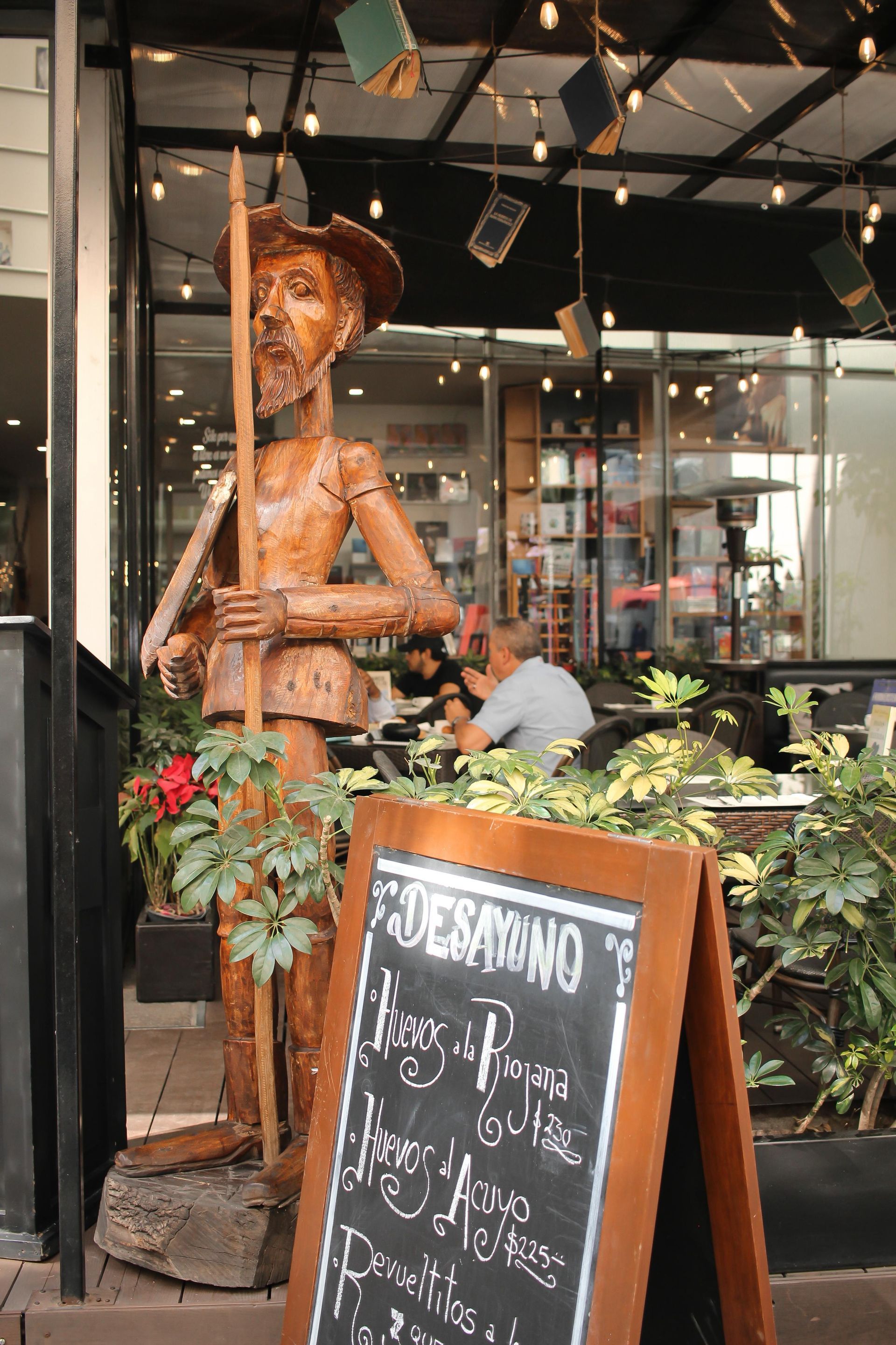 Estatua de vaquero de madera junto a una pizarra con el menú de una cafetería en un local lleno de plantas.