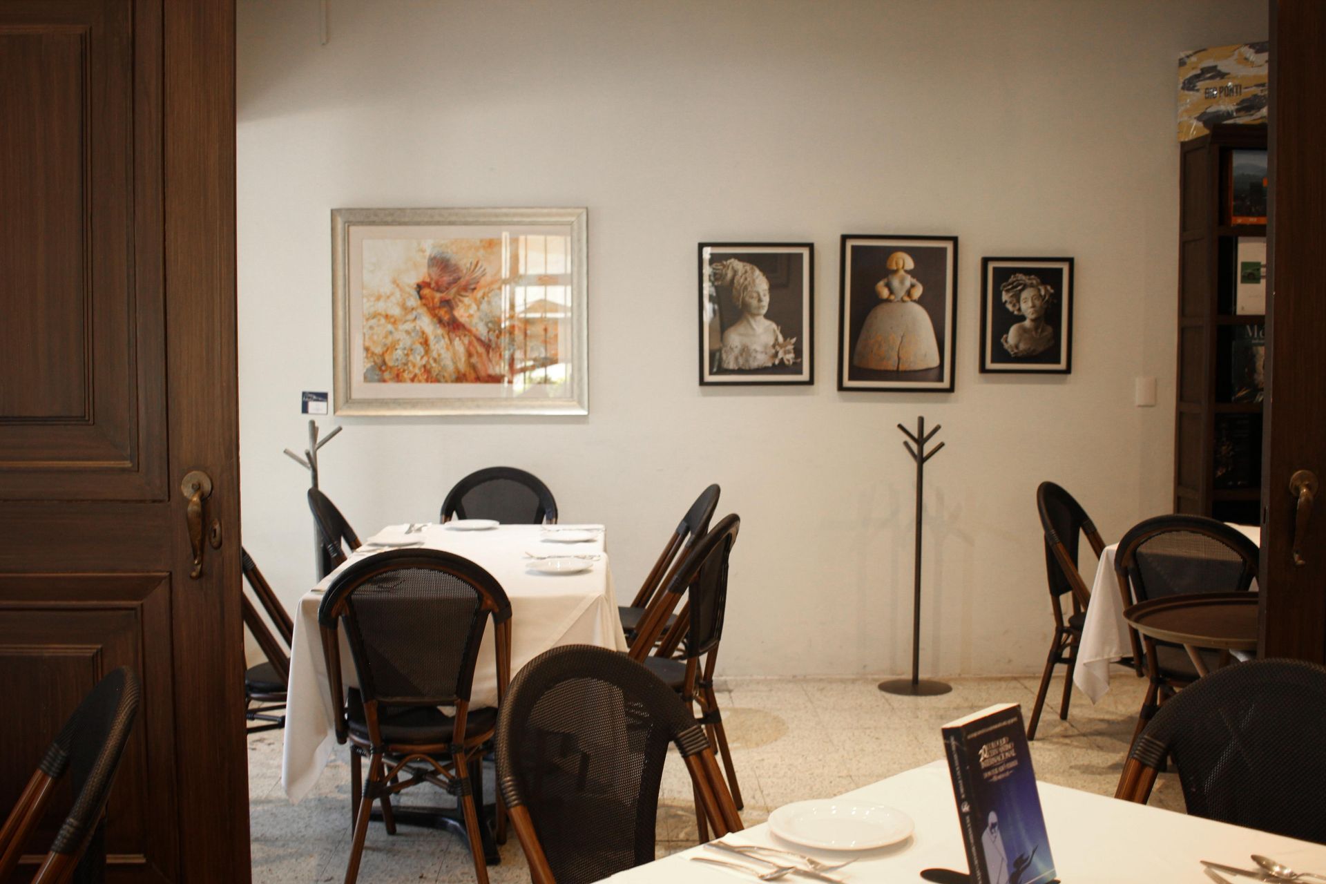 Elegante comedor con paredes blancas, cuadros enmarcados, mesas redondas y sillas de mimbre oscuro.