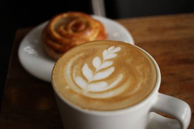 Un café con leche decorado con hojas, junto a un pastel sobre una mesa de madera.