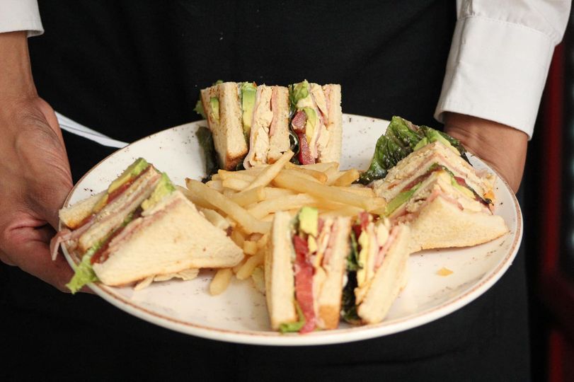 Bandeja de sándwiches club variados en un plato sostenido por un camarero con uniforme negro.