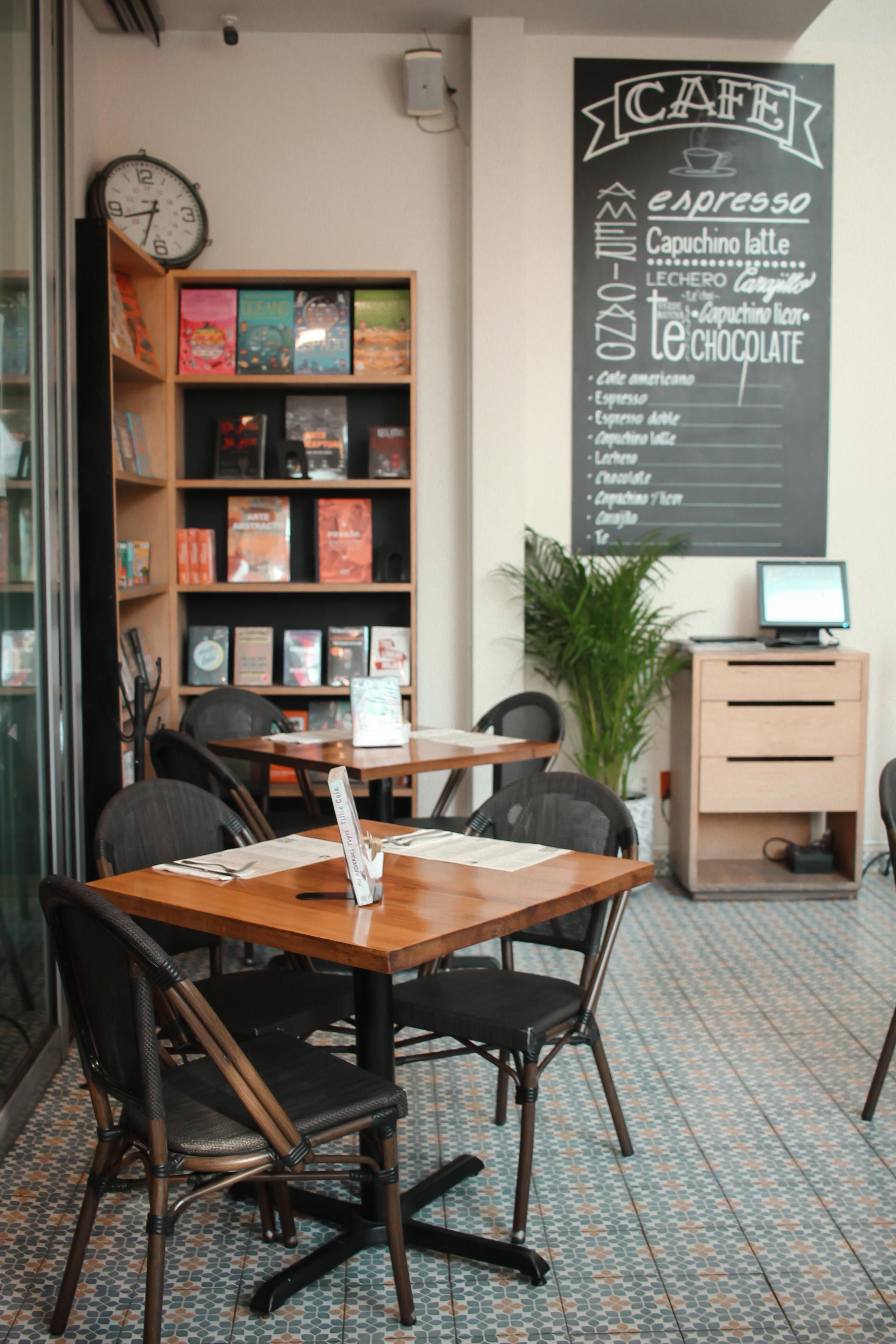 Interior acogedor de una cafetería con mesas de madera, estantería, pizarra con el menú y una pequeña planta en maceta.