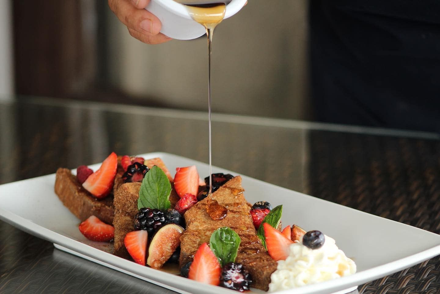 Fresas y frutos rojos bañados en almíbar sobre un plato blanco, acompañados de crema batida y una porción de pastel de chocolate.