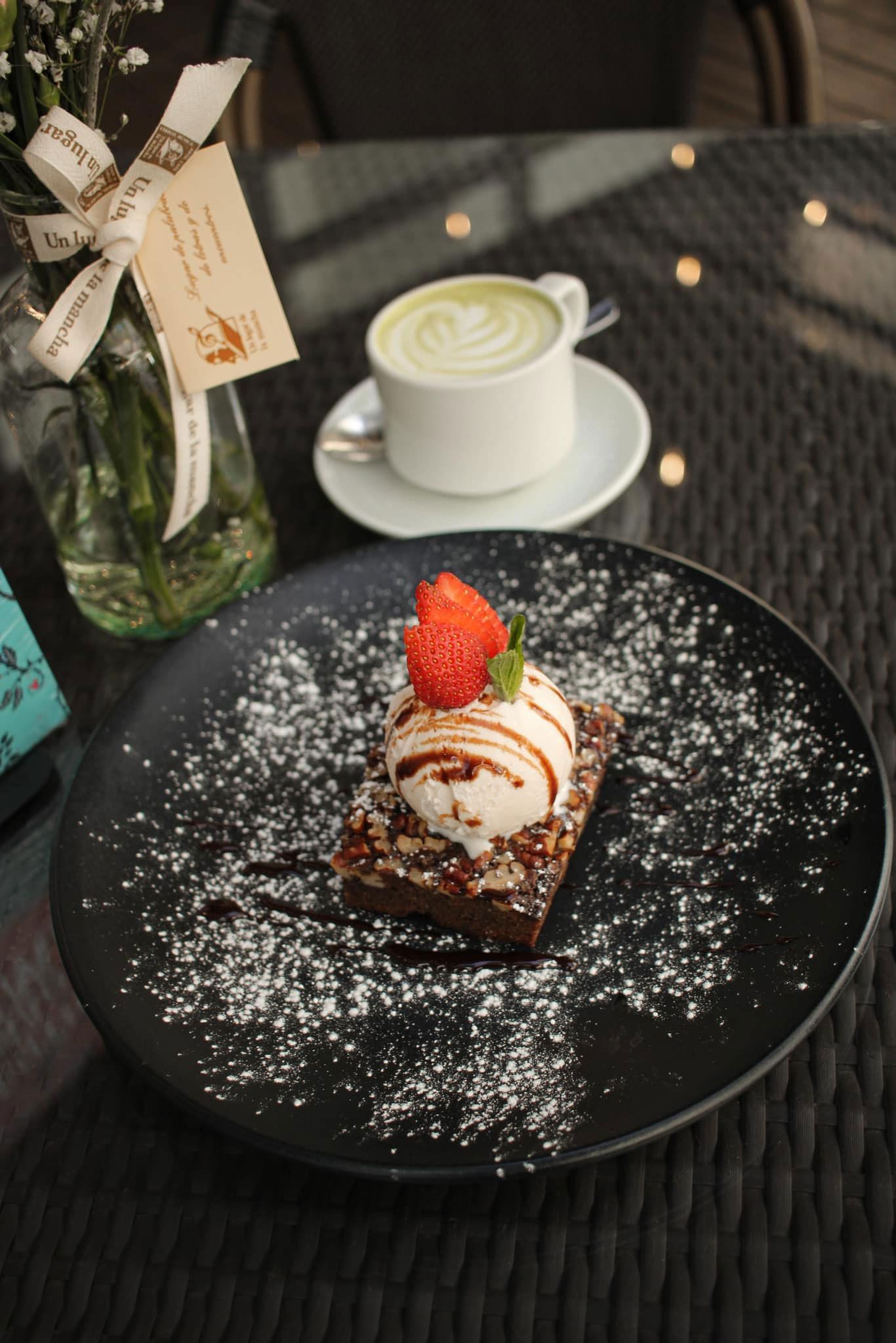 Postre de chocolate en un plato negro con azúcar glas y adorno de fresa, junto a una taza de café sobre una mesa.