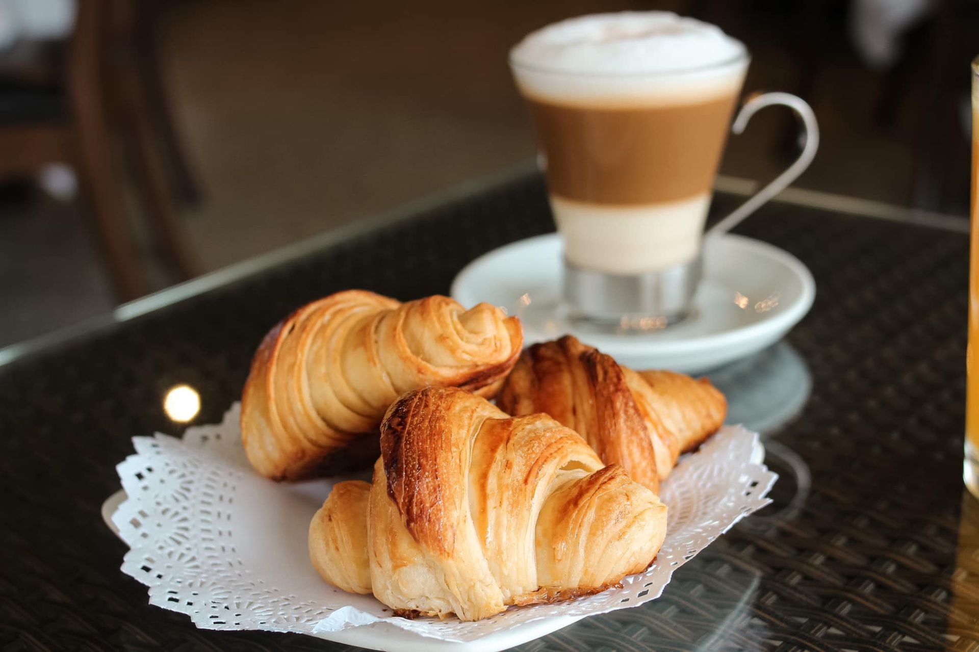 Croissants sobre un tapete de encaje al lado de un café con leche en una taza de cristal sobre la mesa de un café.