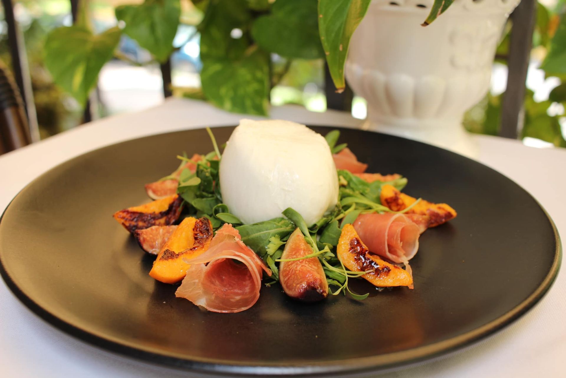 Aperitivo gourmet en un plato negro con embutido, verduras y una bola de helado blanco tipo sorbete en un ambiente de restaurante.