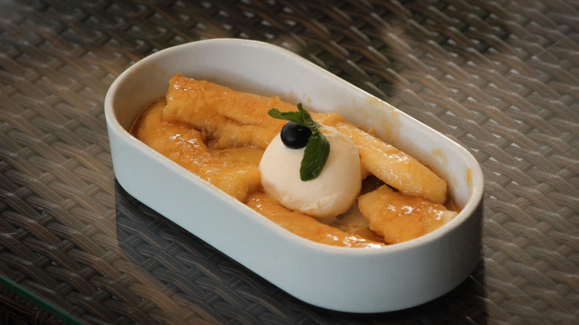 Postre en un plato blanco con salsa de naranja, una bola de helado y guarnición de menta sobre una mesa texturizada.