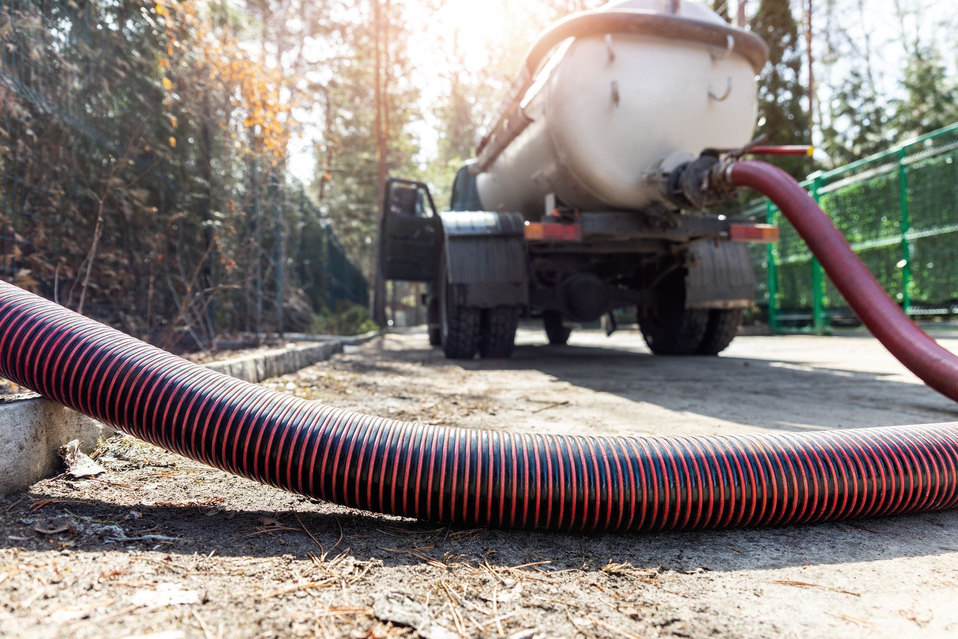 Septic truck with large hoses pumping waste from an underground tank in a wooded area.