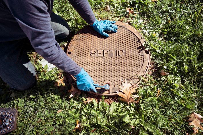 Person in blue gloves opens a septic tank cover in a grassy area.