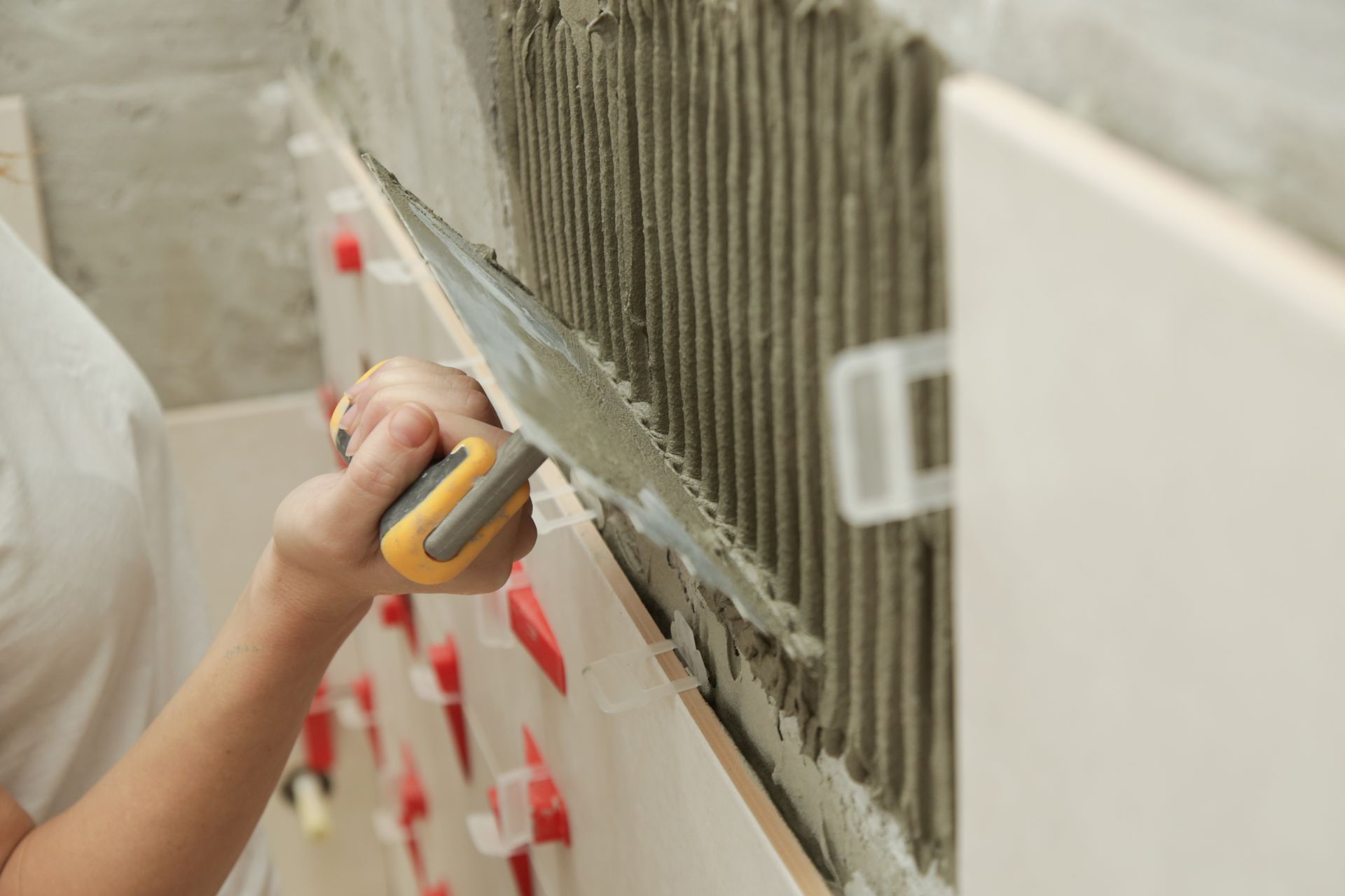 Person applying gray adhesive to wall with a notched trowel during tile installation