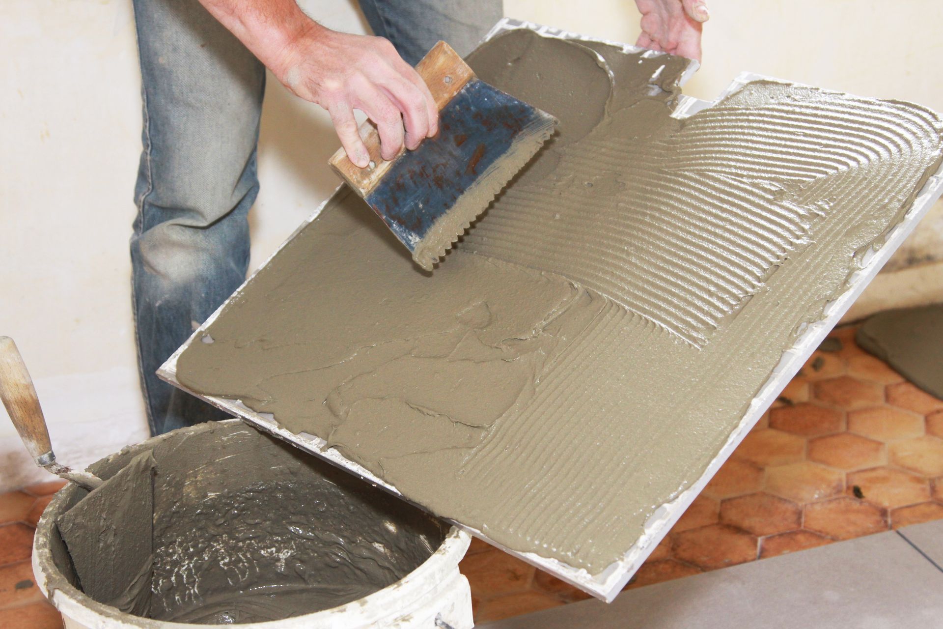 Person spreading gray mortar on a tile backer board with a trowel beside a bucket.