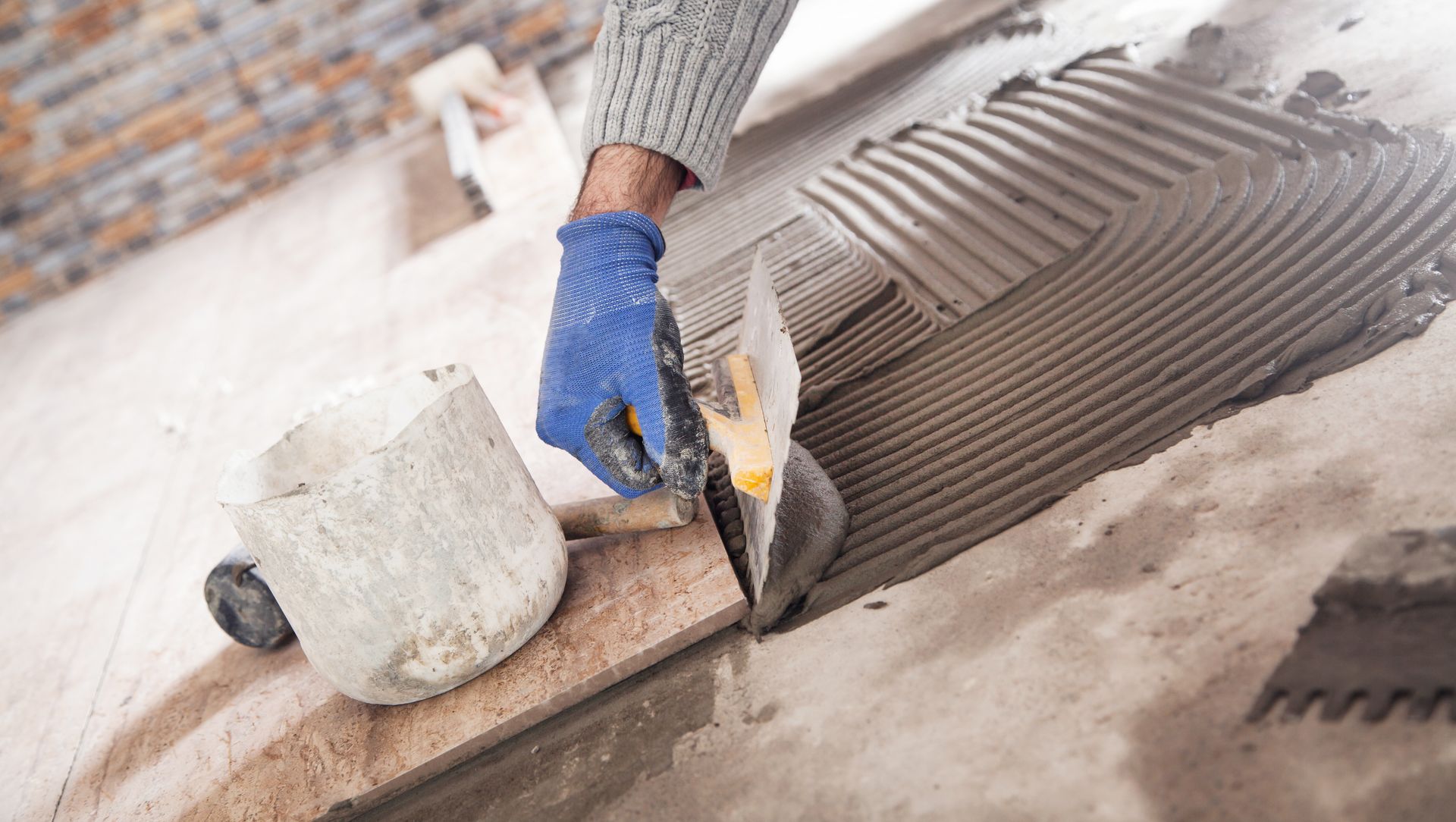 Gloved worker spreading adhesive with a trowel beside a bucket on a floor under construction