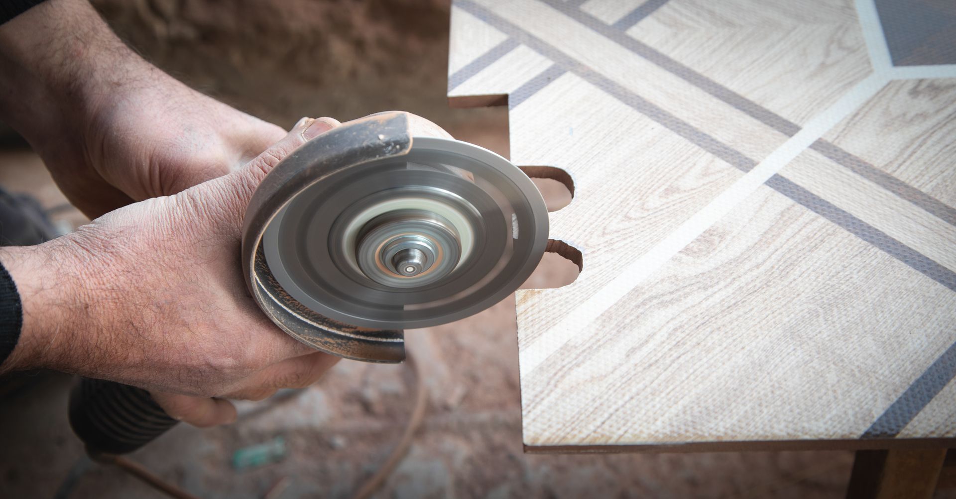 Hands holding a circular metal tool part over a tiled floor with cardboard packaging in the background