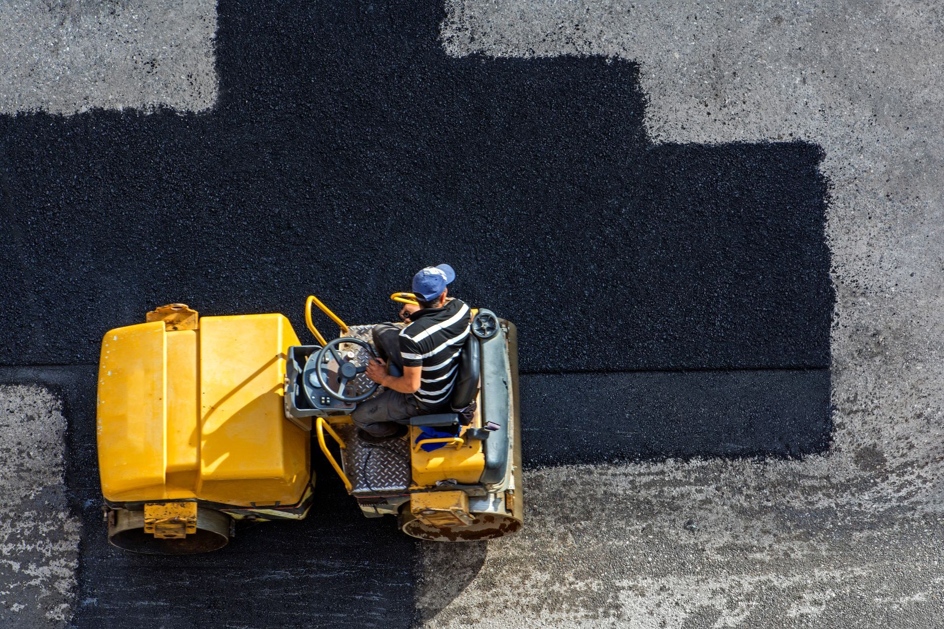 An aerial view of a worker operating a yellow steamroller to pave a dark asphalt surface onto a grey road.