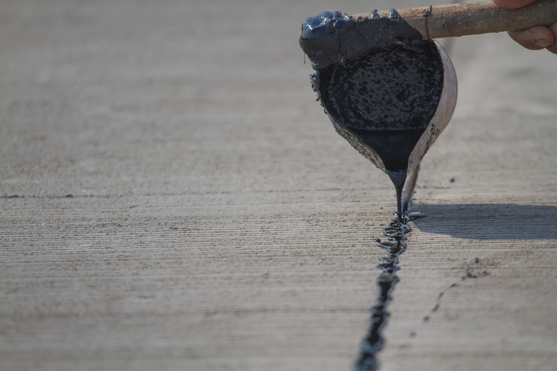 A worker pouring black sealant from a metal ladle into a crack in a concrete surface.