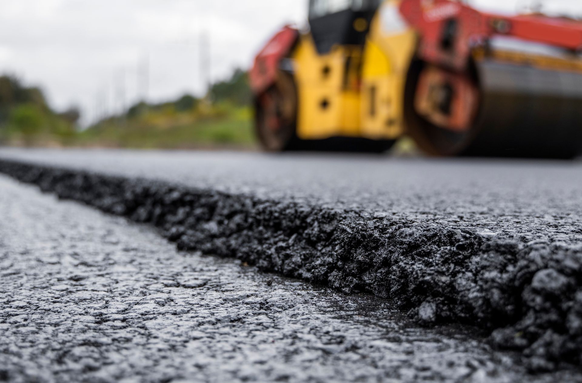A freshly laid asphalt road surface in the foreground with a yellow and red road roller blurred in the background.