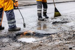 Road workers in high-visibility orange clothing use a heating torch and broom to repair a pothole in a wet street.