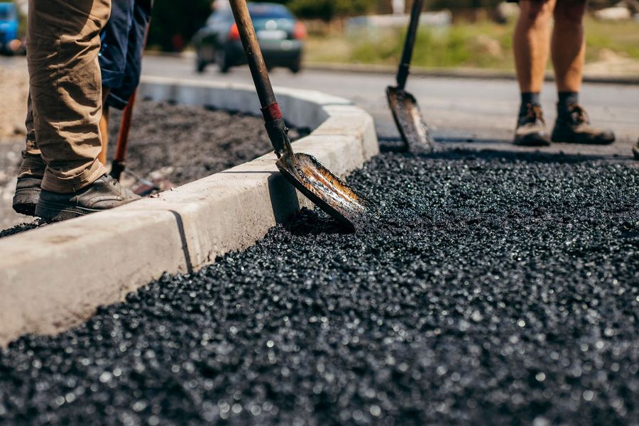 Construction workers use shovels to smooth fresh black asphalt along a concrete curb on a sunny day.