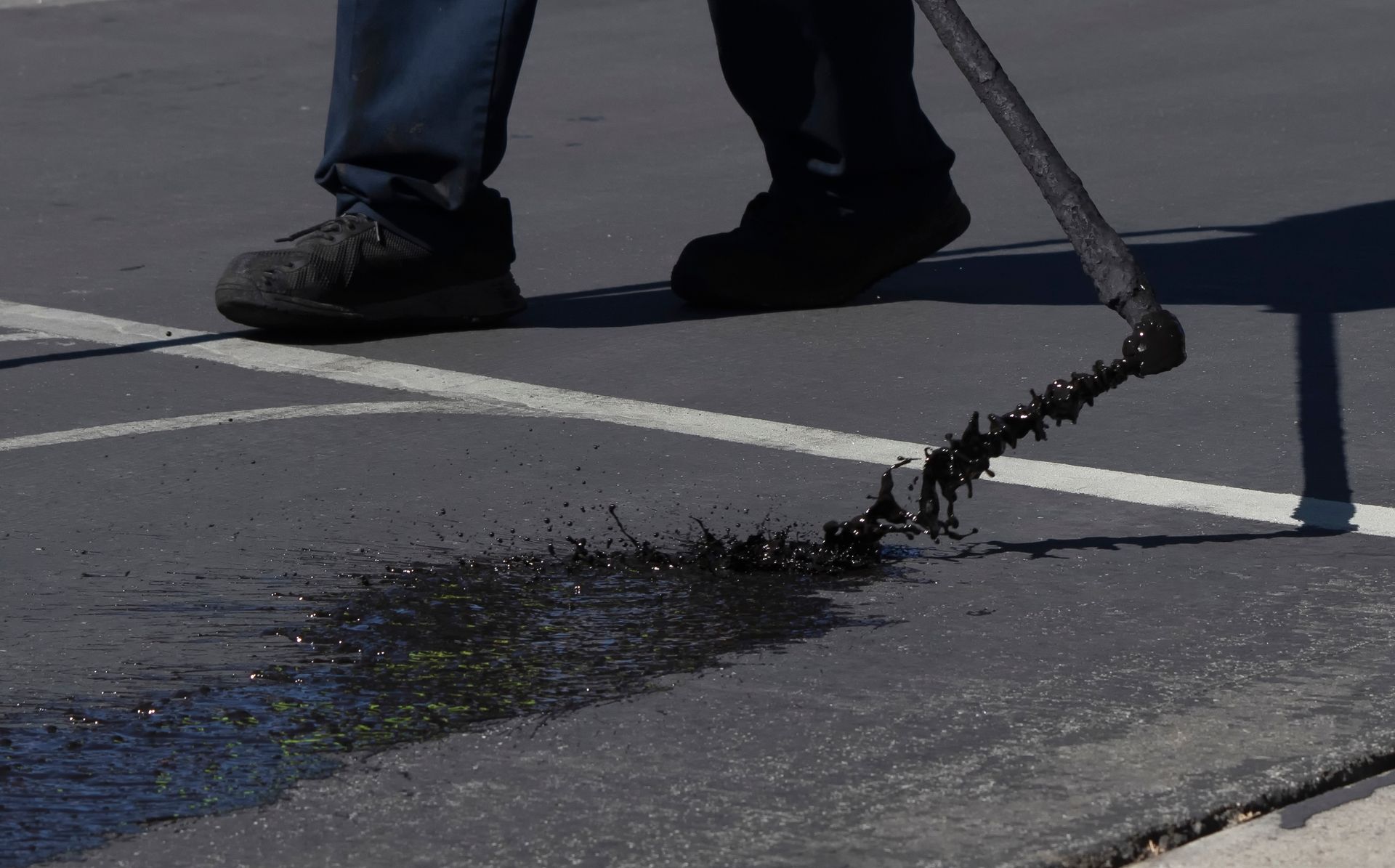 A worker in dark pants and shoes uses a long-handled tool to spray black asphalt sealant onto a paved surface.