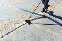 A person uses a crack-filling tool to apply black sealant over yellowed cracks in grey asphalt.