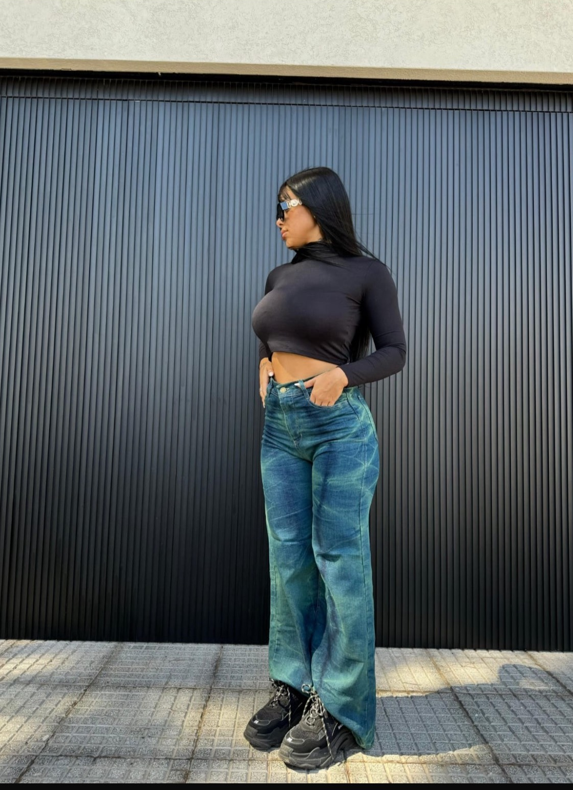 Una mujer que viste un top negro y jeans azules está parada frente a la puerta de un garaje.