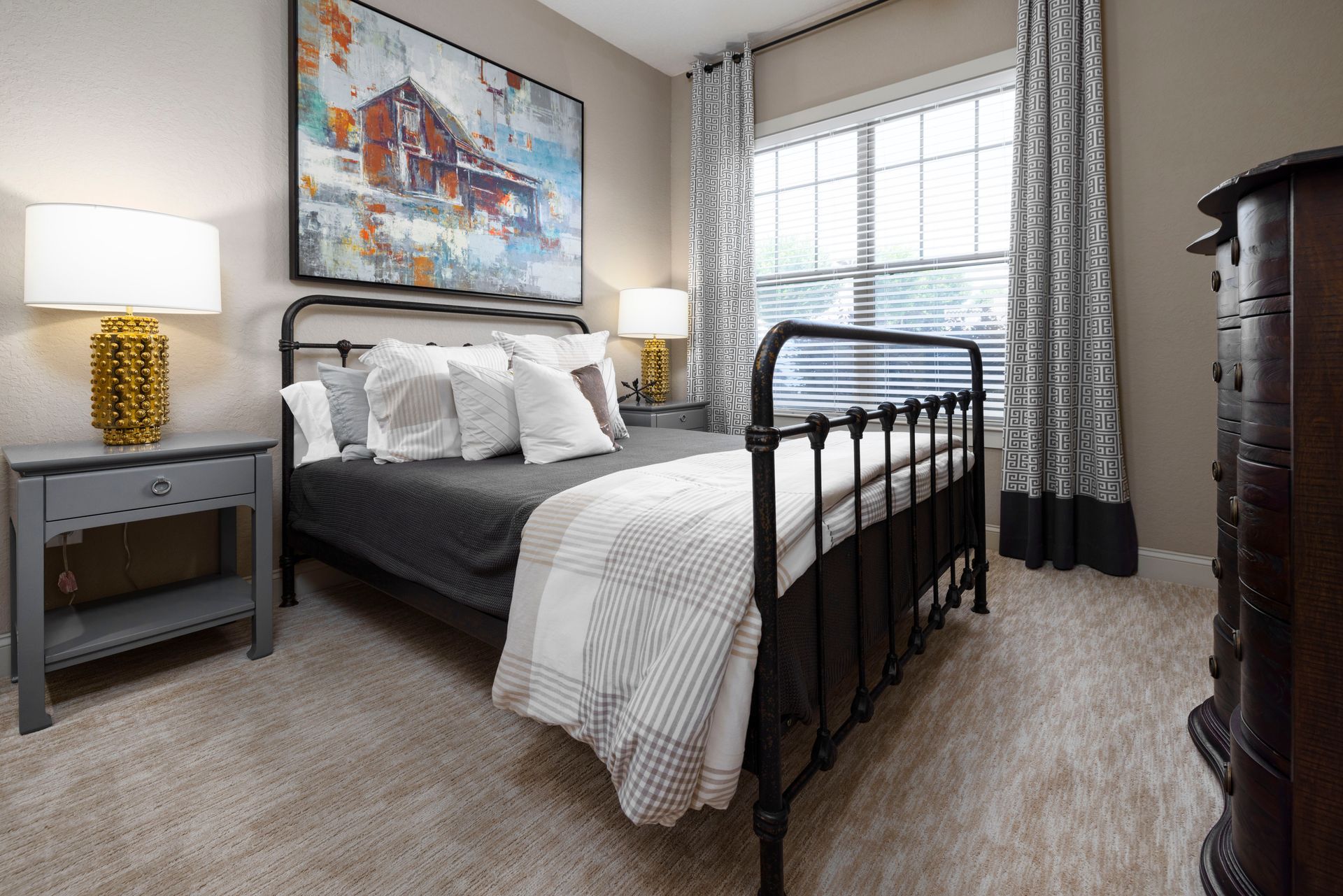 A bedroom with a black metal bed frame, grey nightstand with a gold lamp, patterned curtains, and an abstract painting.
