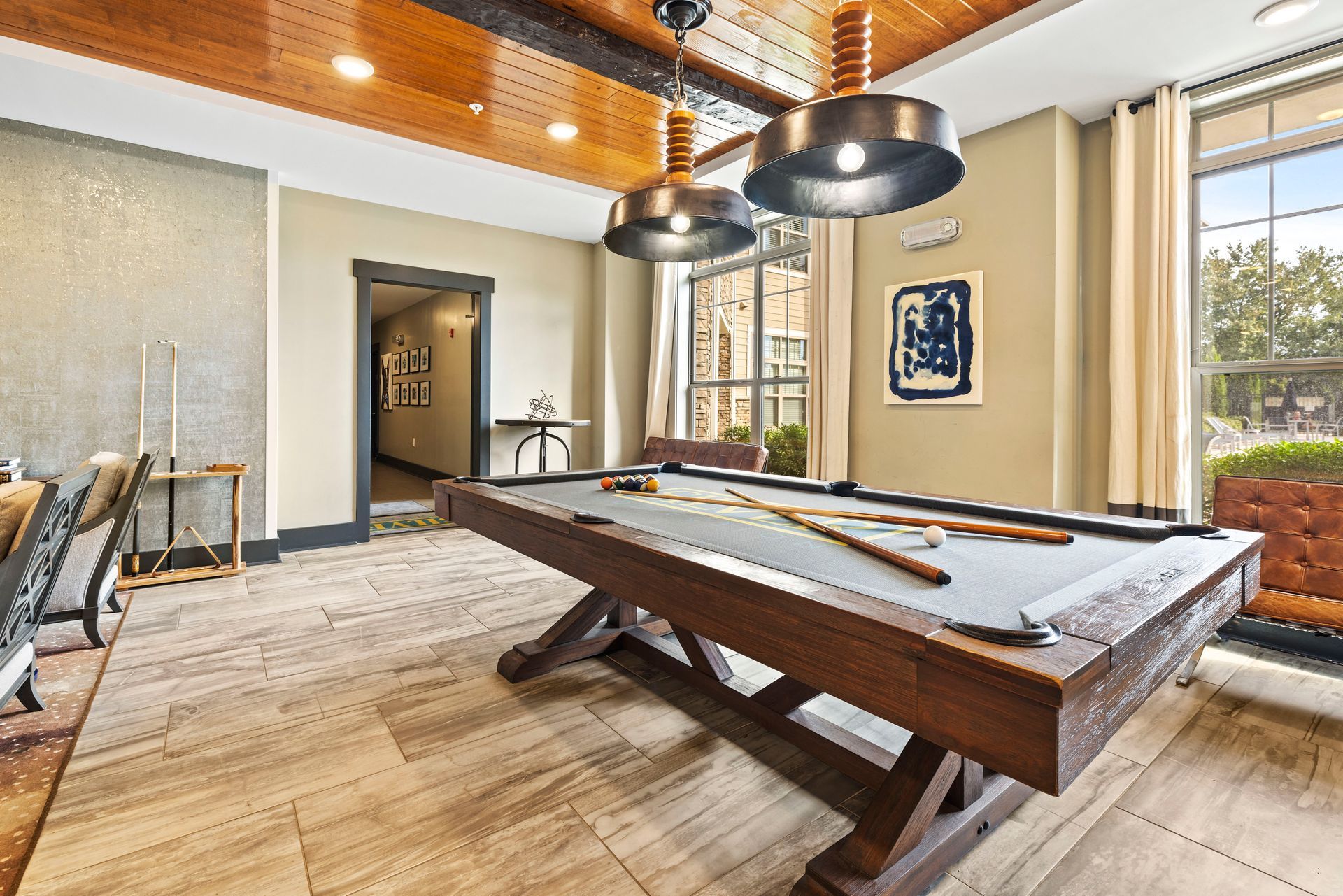 A pool table in a lounge area with a wooden ceiling and large windows. The room has neutral colors and modern lighting.