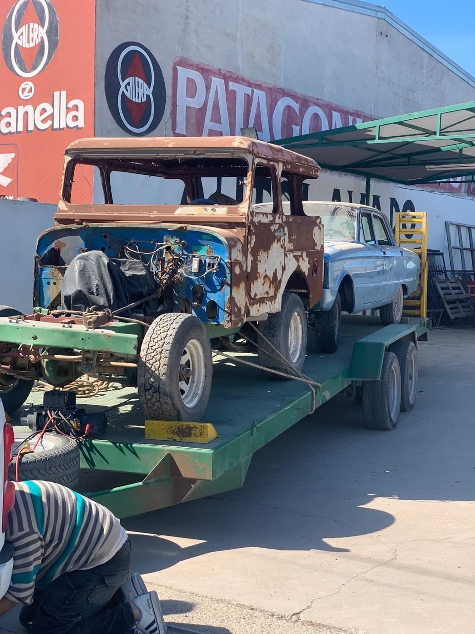 Camioneta antigua oxidada atada a un remolque de plataforma frente a una tienda.