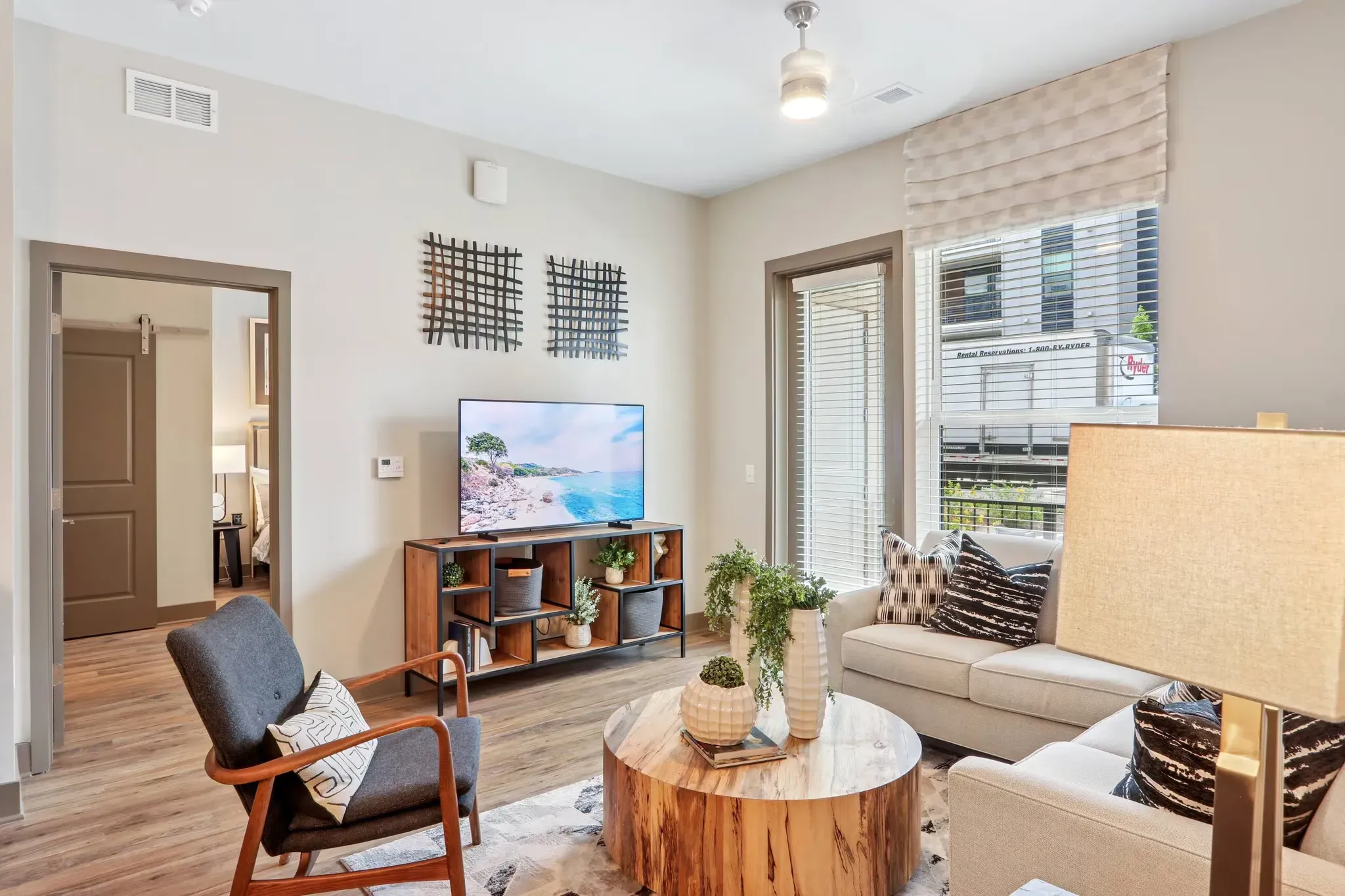Living room in a modern apartment with a sofa, armchair, round wood coffee table, TV stand, and large window with blinds.