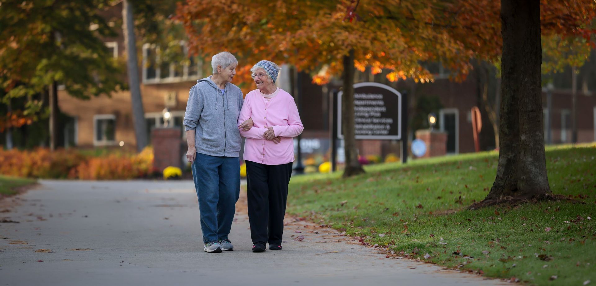 Two people walking arm in arm on a tree-lined path in autumn, with fallen leaves and a park sign nearby