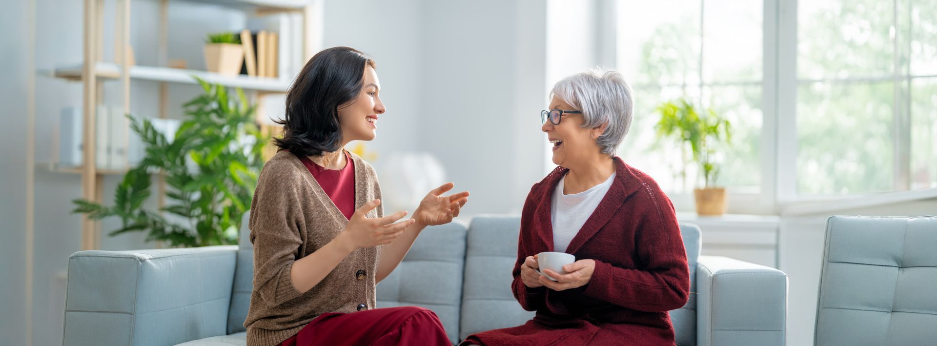 Two people chatting on a couch in a bright living room, holding cups and smiling