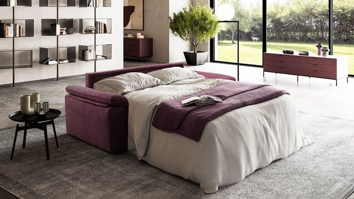 Camera da letto moderna con letto bianco, biancheria da letto color prugna, mensole a vista e ampie finestre che si affacciano sul verde.