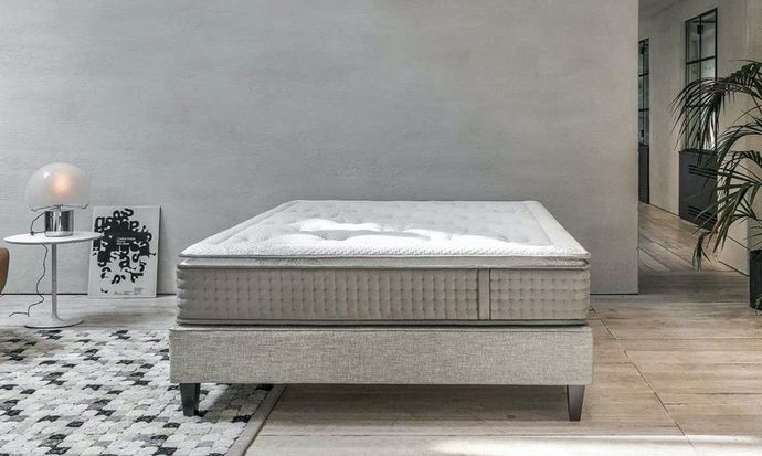 Letto moderno con testiera trapuntata grigio chiaro in una camera da letto minimalista, vicino a una pianta e un comodino.