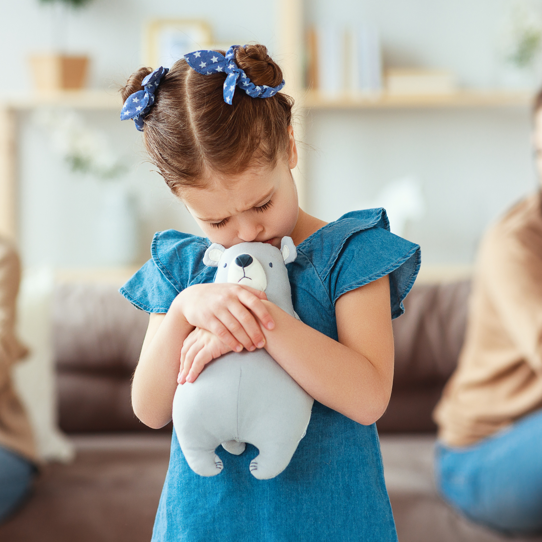 A person with curly hair and blue bows holds a stuffed animal to their forehead, appearing upset in a living room.