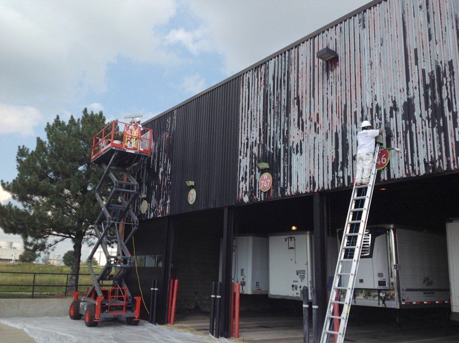 Workers painting a weathered black building exterior. One uses a lift, the other a ladder.