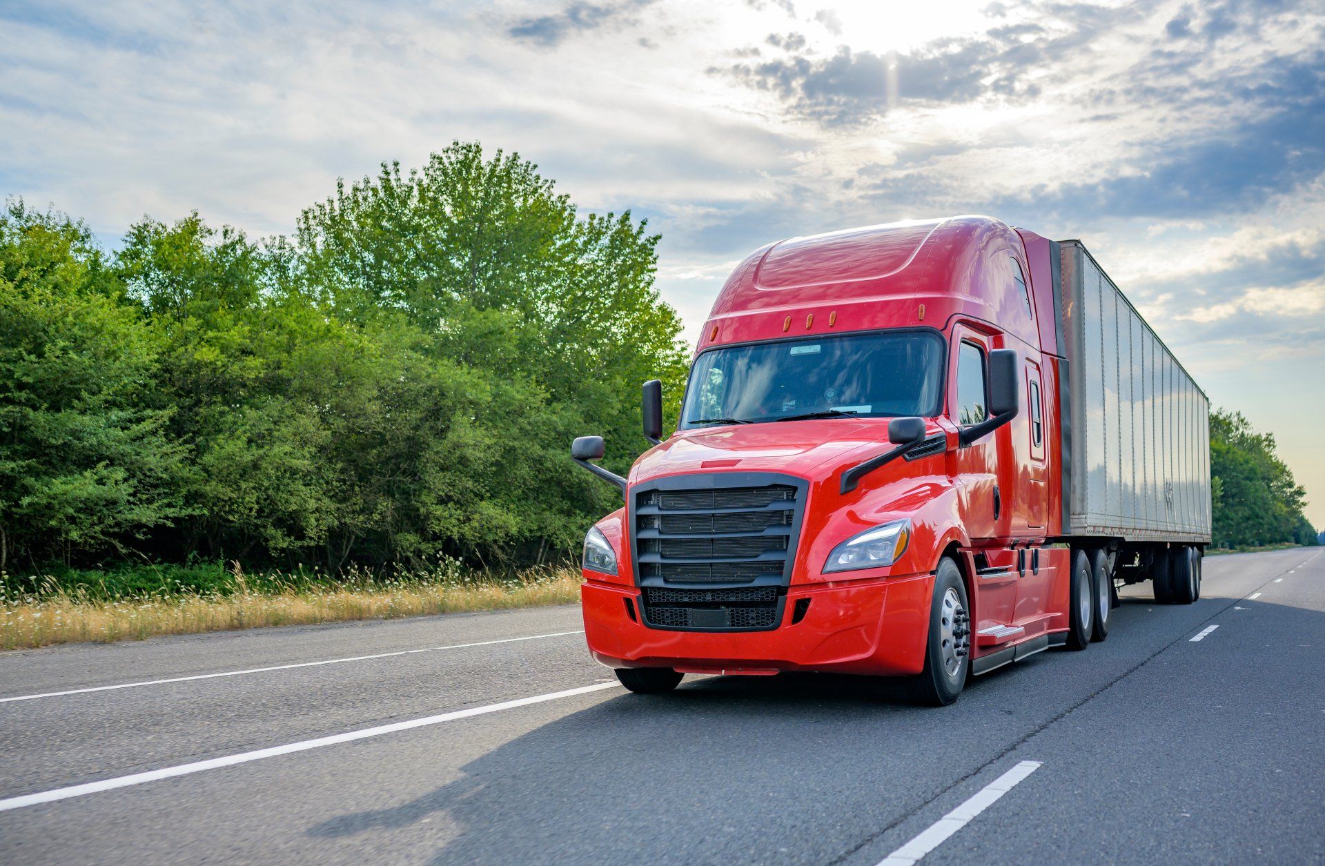 Red semi-truck on highway, driving past green trees under a cloudy sky.