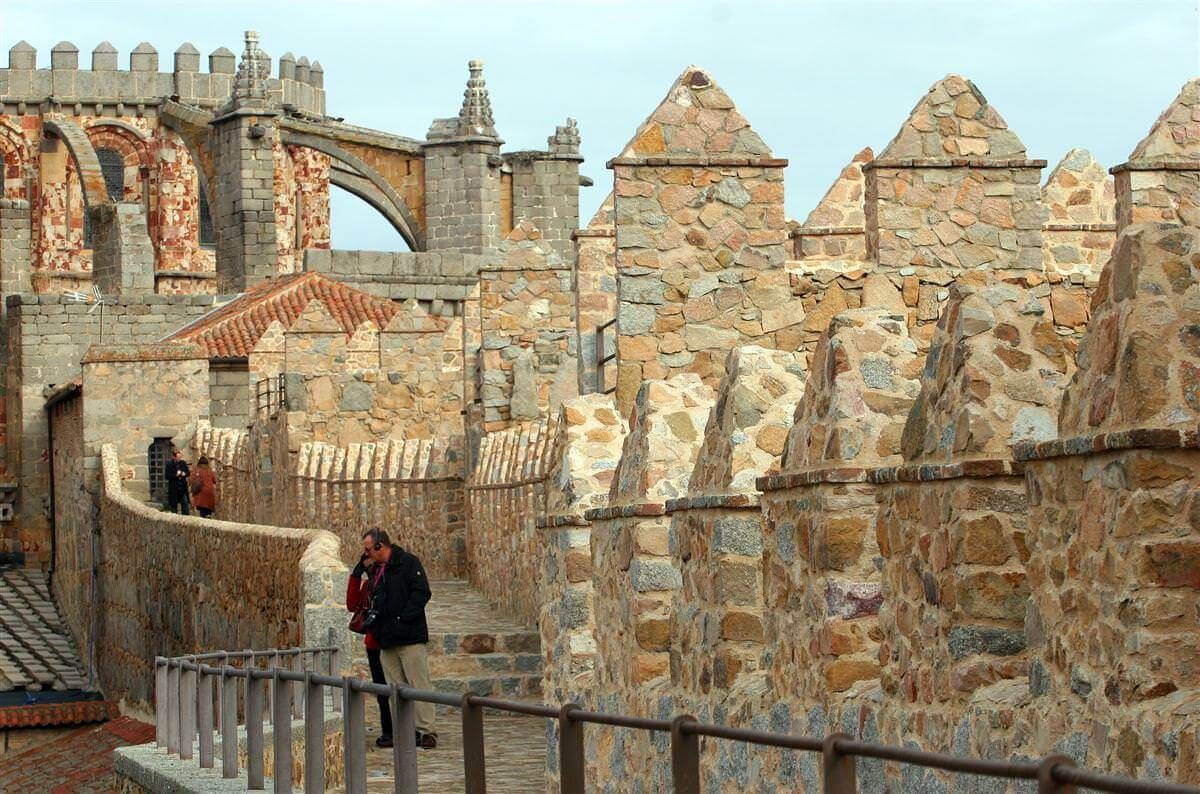 Murallas de piedra del castillo con un pasillo abovedado, tejados de tejas rojas y algunos visitantes paseando por el sendero.