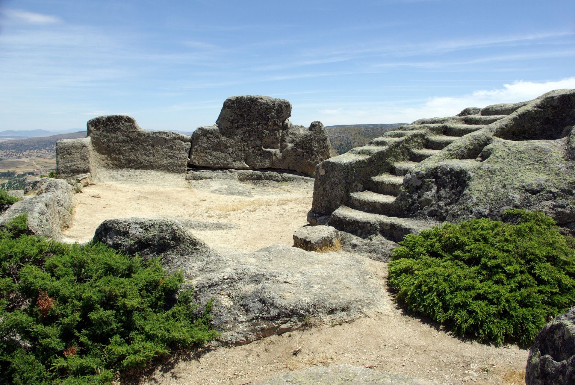 Ruinas cubiertas de hierba de muros bajos de piedra en un paisaje boscoso bajo un cielo parcialmente nublado.