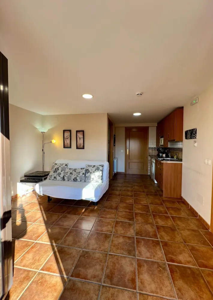 Sala de estar con suelo de baldosas marrones, sofá blanco y cocina abierta al fondo.