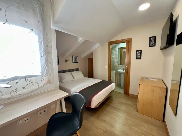 Habitación pequeña con cama, escritorio junto a la ventana, silla y puerta de baño bajo un techo inclinado.